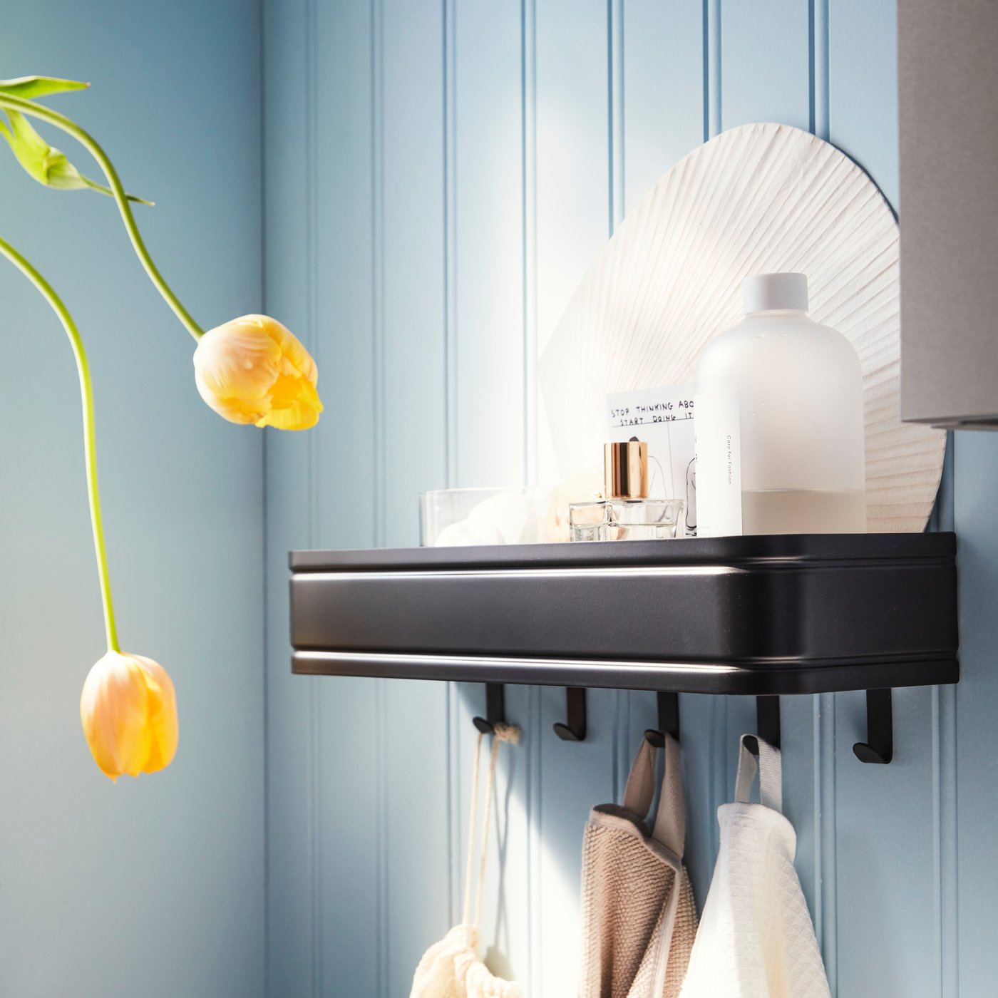 A black LILLASJÖN wall shelf with five hooks is mounted on a blue wall, with products on the shelf and towels on the hooks.