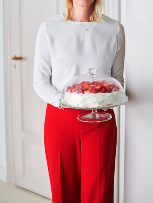 Camera de zi cu perete alb, usa, o persoana cu bluza alba si pantaloni rosii, tinand in mana un stand de servire cu capac, cu prajitura.