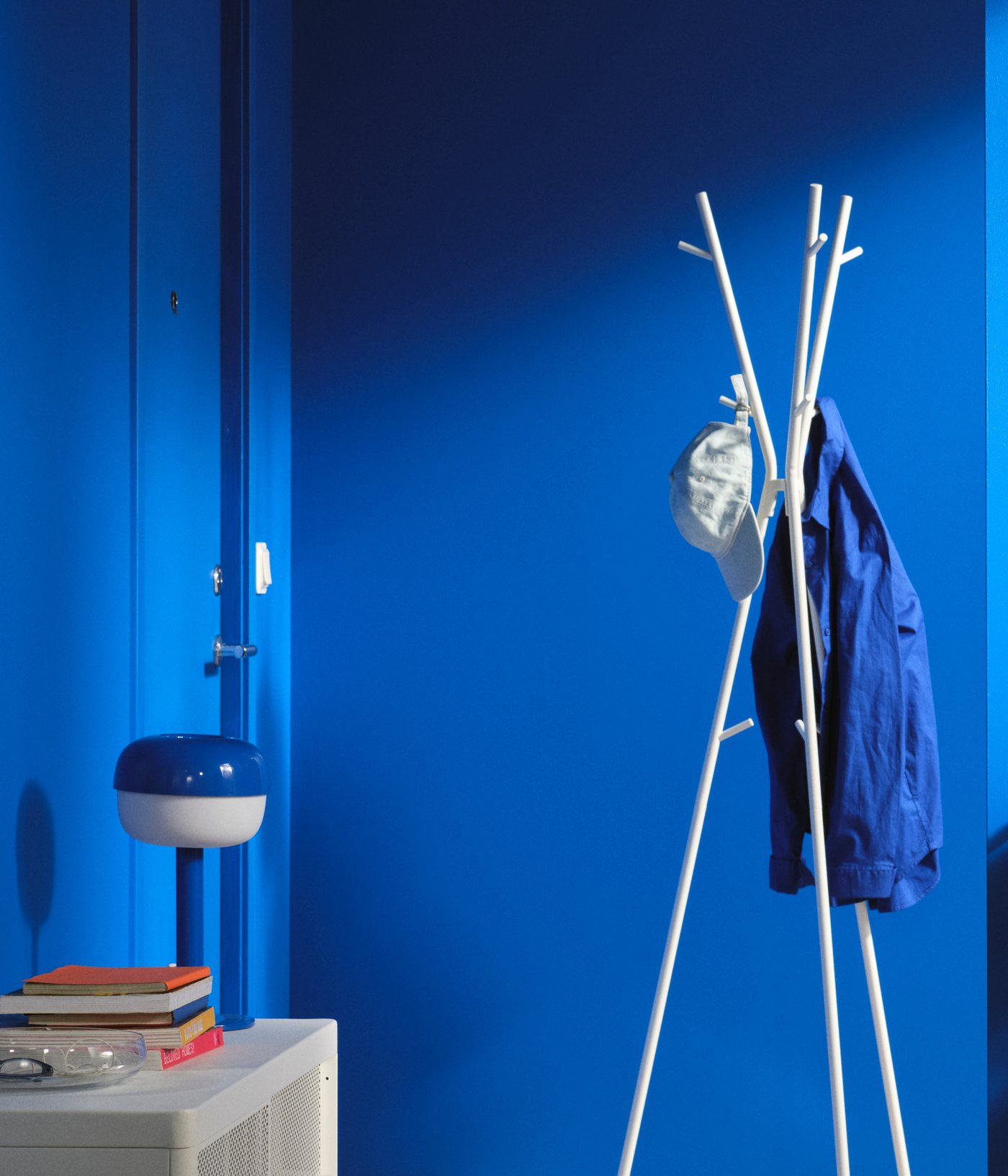 A blue hallway with an EKRAR hat/coat stand in the centre holding a coat and baseball cap, beside a white NISSAFORS trolley.