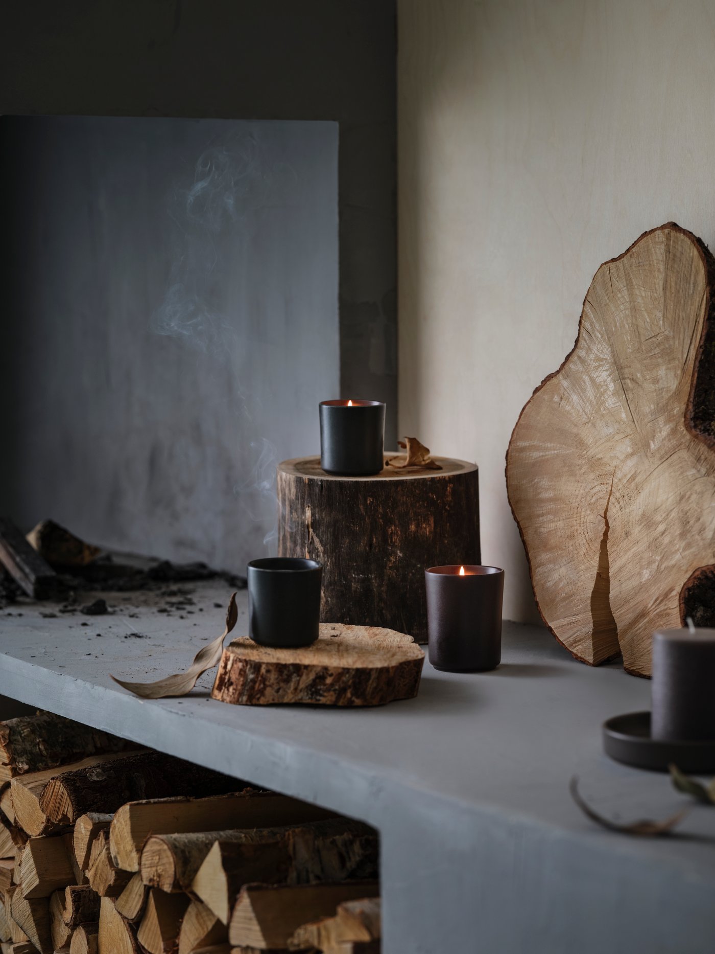 Tre candele profumate BEHJÄRTAD in vasi di ceramica, appoggiate su ceppi di legno su un camino in pietra rustica.