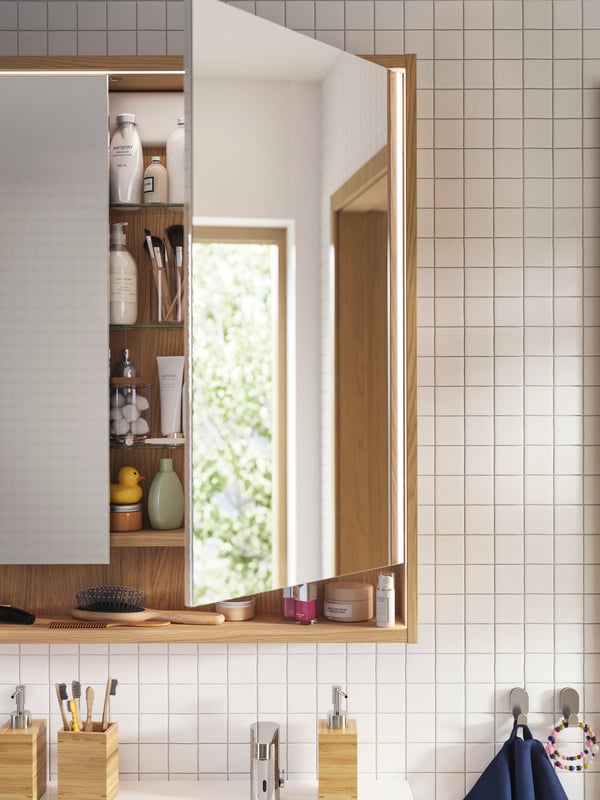 Une armoire à pharmacie FAXÄLVEN en chêne avec éclairage intégré, installé au-dessus d’un lavabo, dans une salle de bains avec carrelage blanc.