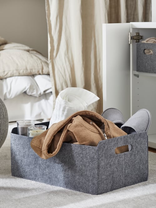 A box containing a pair of grey slippers, scented candles and clothing items is placed next to a pair of beige curtains.