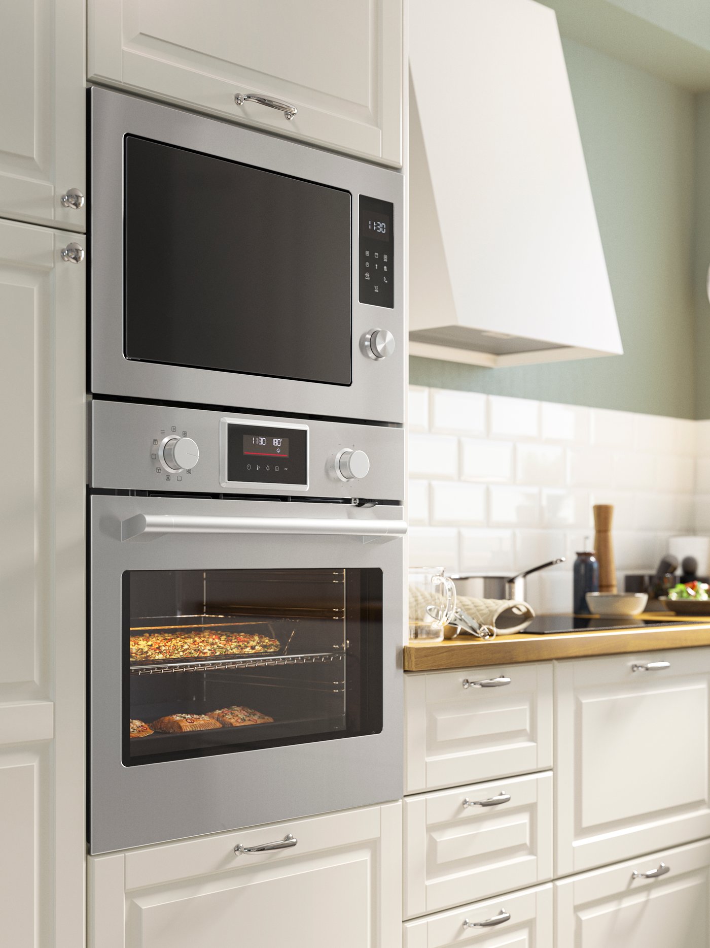 Cucina con mobili bianchi e piano di lavoro in legno chiaro. A sinistra, una colonna forno con un microonde in acciaio inossidabile in alto e un forno sottostante con cibo in cottura.