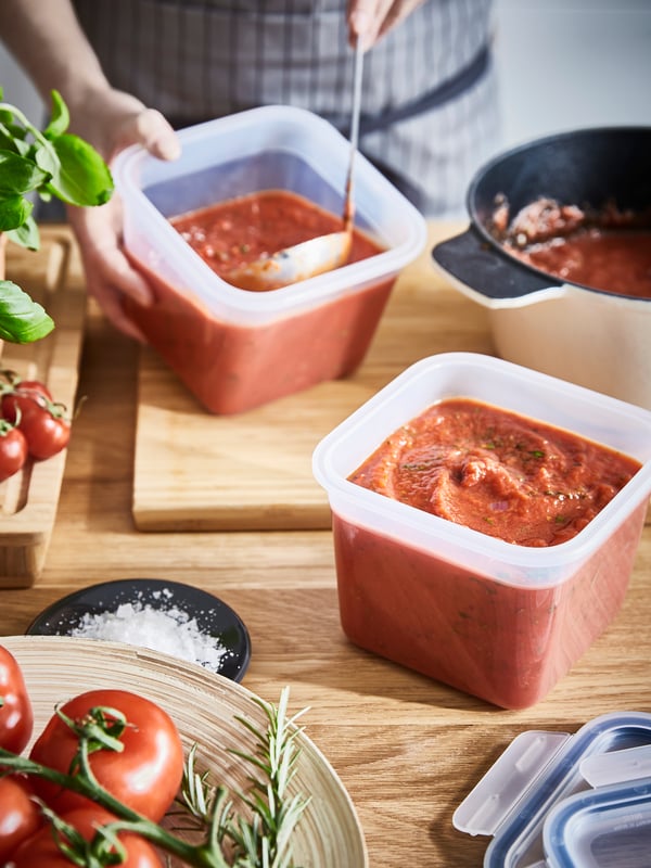 A person wearing a checked apron is spooning tomato sauce from a pan into two plastic food containers.