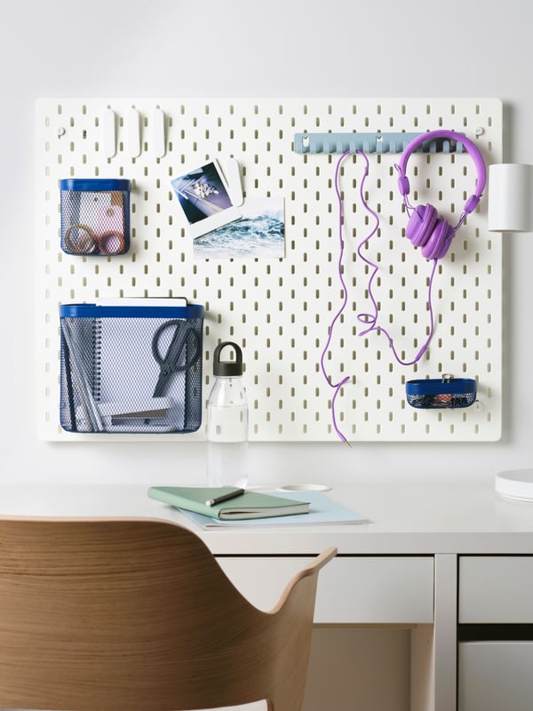 A white memo board hanging above a white MICKE desk with purple headphones and blue baskets clipped to it, along with various pictures hung up