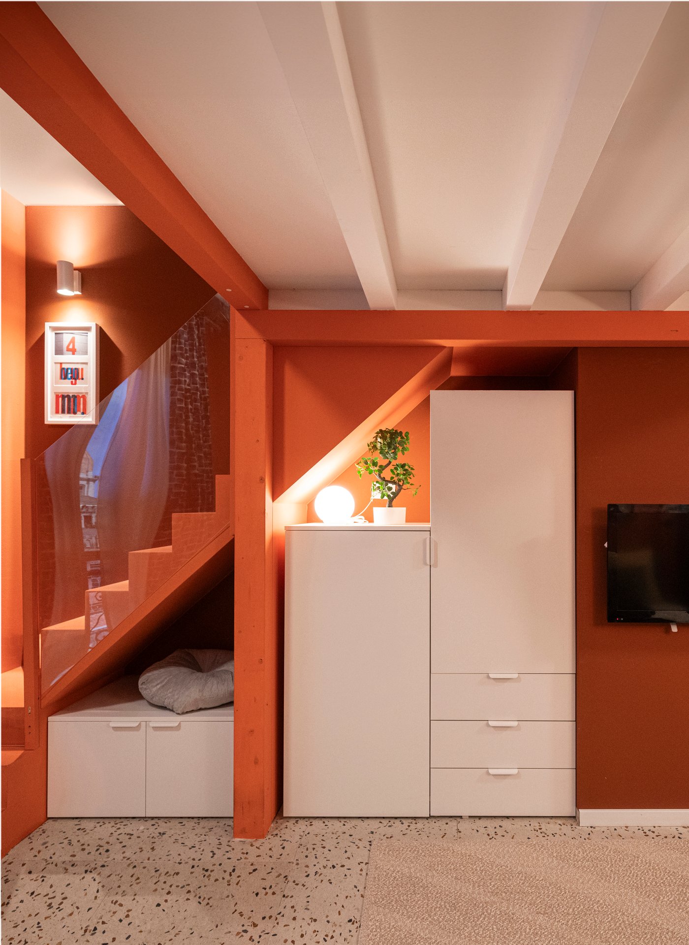 Rum med vit PLATSA garderob under trappa, orange väggar, inbyggd sittplats med kuddar, växt och lampa ovanpå garderoben, samt väggmonterad TV.

