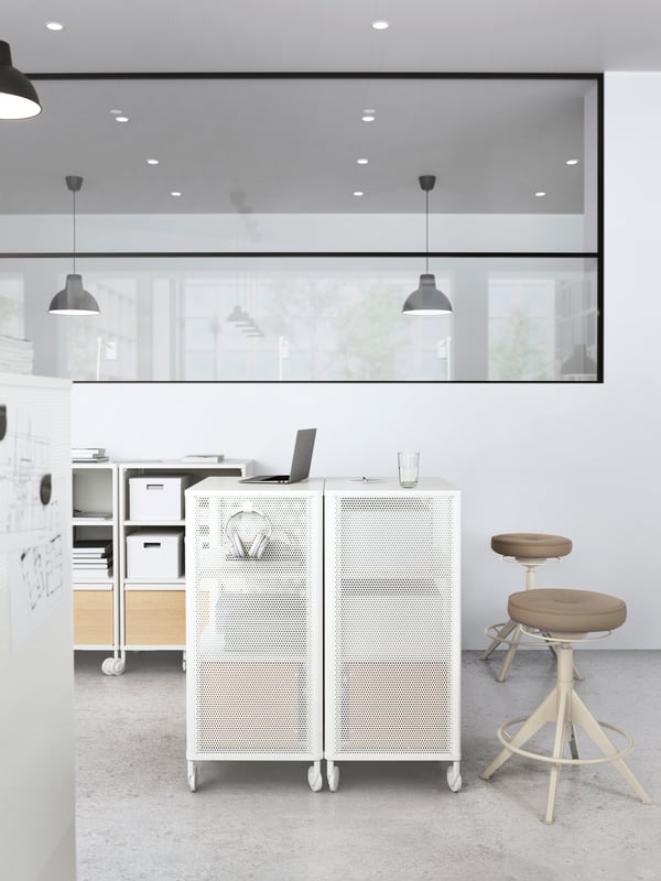 A laptop is sitting on one of two white BEKANT castors in the middle of an office room setting with two beige TROLLBERGET chairs behind.