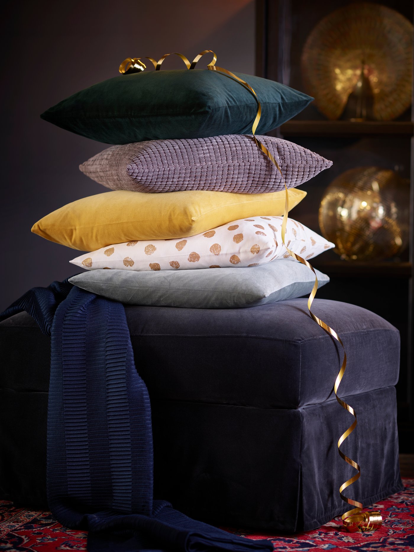 Two corduroy cushions in dark beige and grey-turquoise, and a denim cushion on top of a grey divan bed with padded headboard.