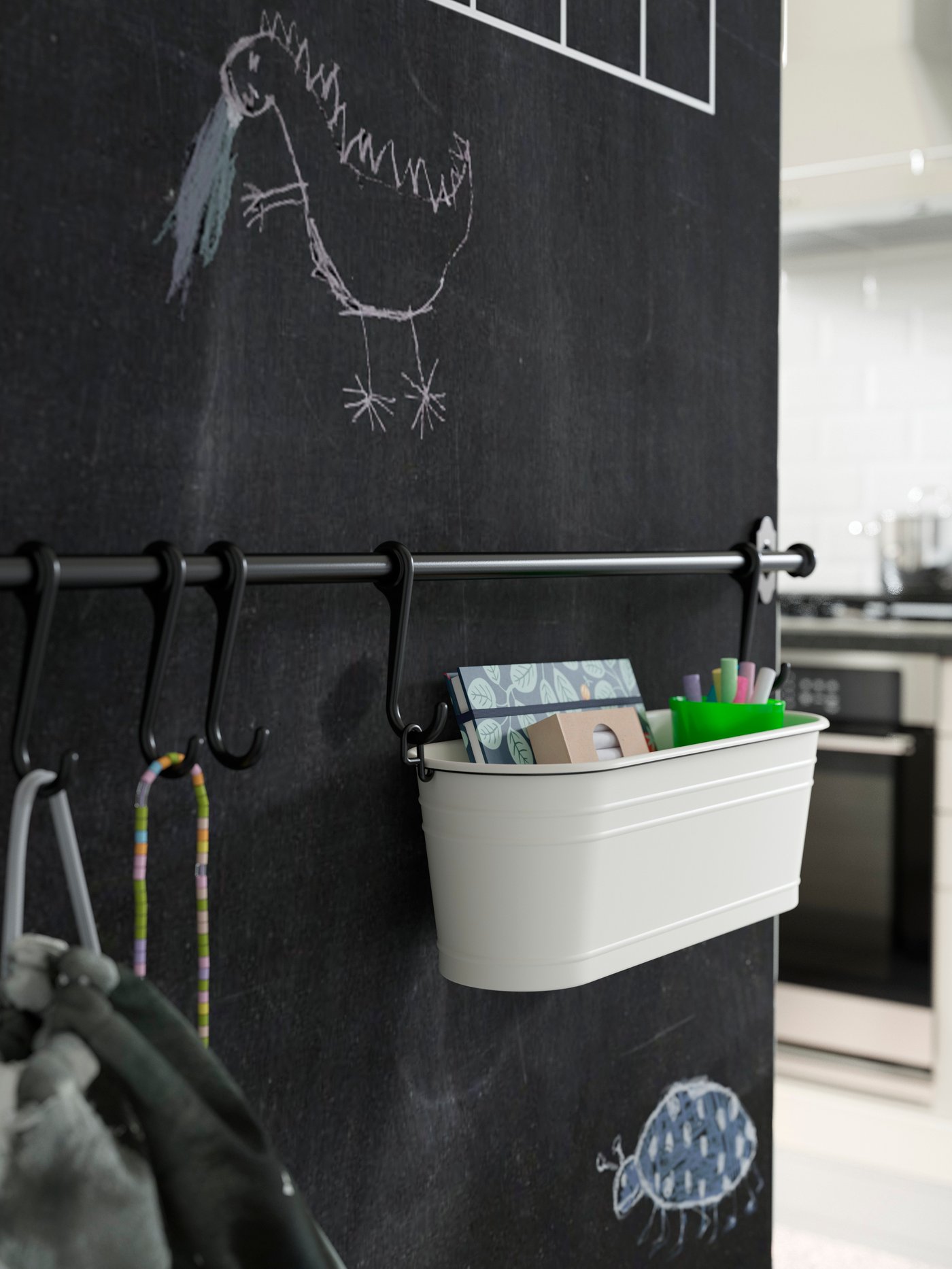 A white condiment box storing a notebook and more hangs from a black rail mounted on a chalkboard wall.