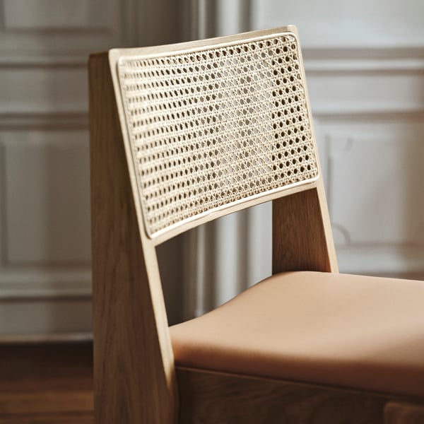 A STOCKHOLM 2025 chair with a solid oak frame, a leather seat, and a rattan backrest stands on parquet flooring.