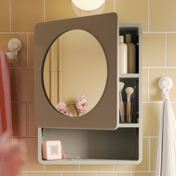 A green IVÖSJÖN mirror cabinet with one door that has a circular mirror on it and a built-in shelf is on a yellow-tiled wall.