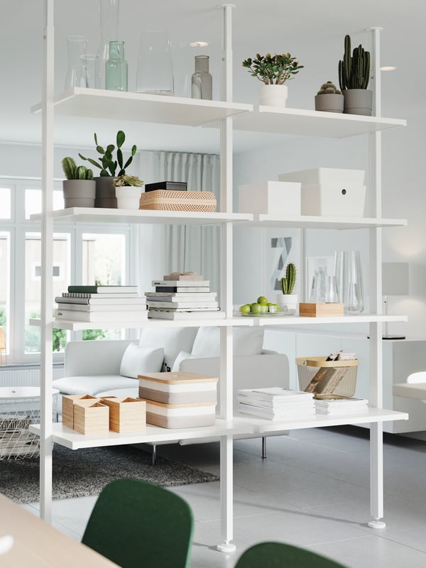 A white storage system that’s used as a room divider. Decorative items, plants, books and storage boxes stand on the shelves.
