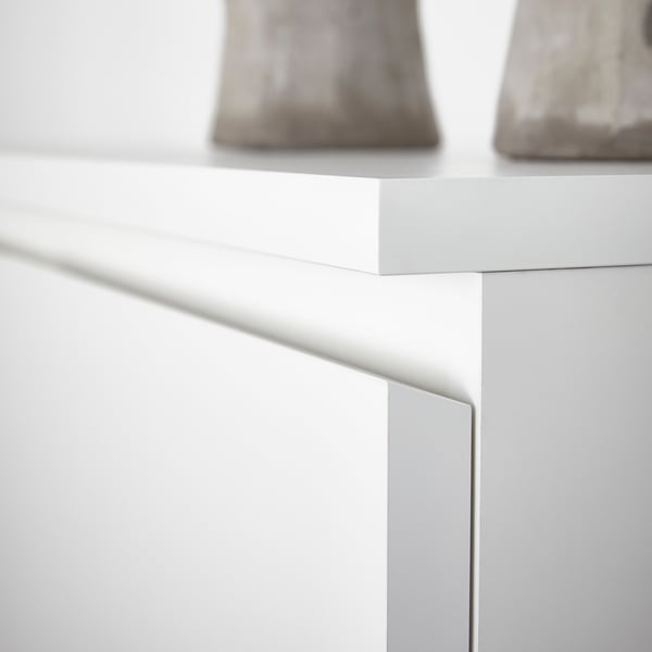 The corner of the surface and the top drawer of a white MALM chest with 2 drawers with two light grey objects on top of it.