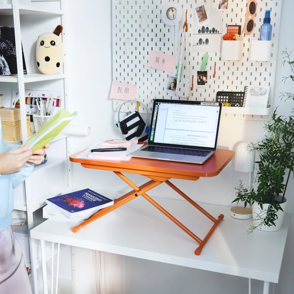 The orange DUBBLA laptop stand with a laptop placed on a white desk in front of a white pegboard and a small pink mirror.