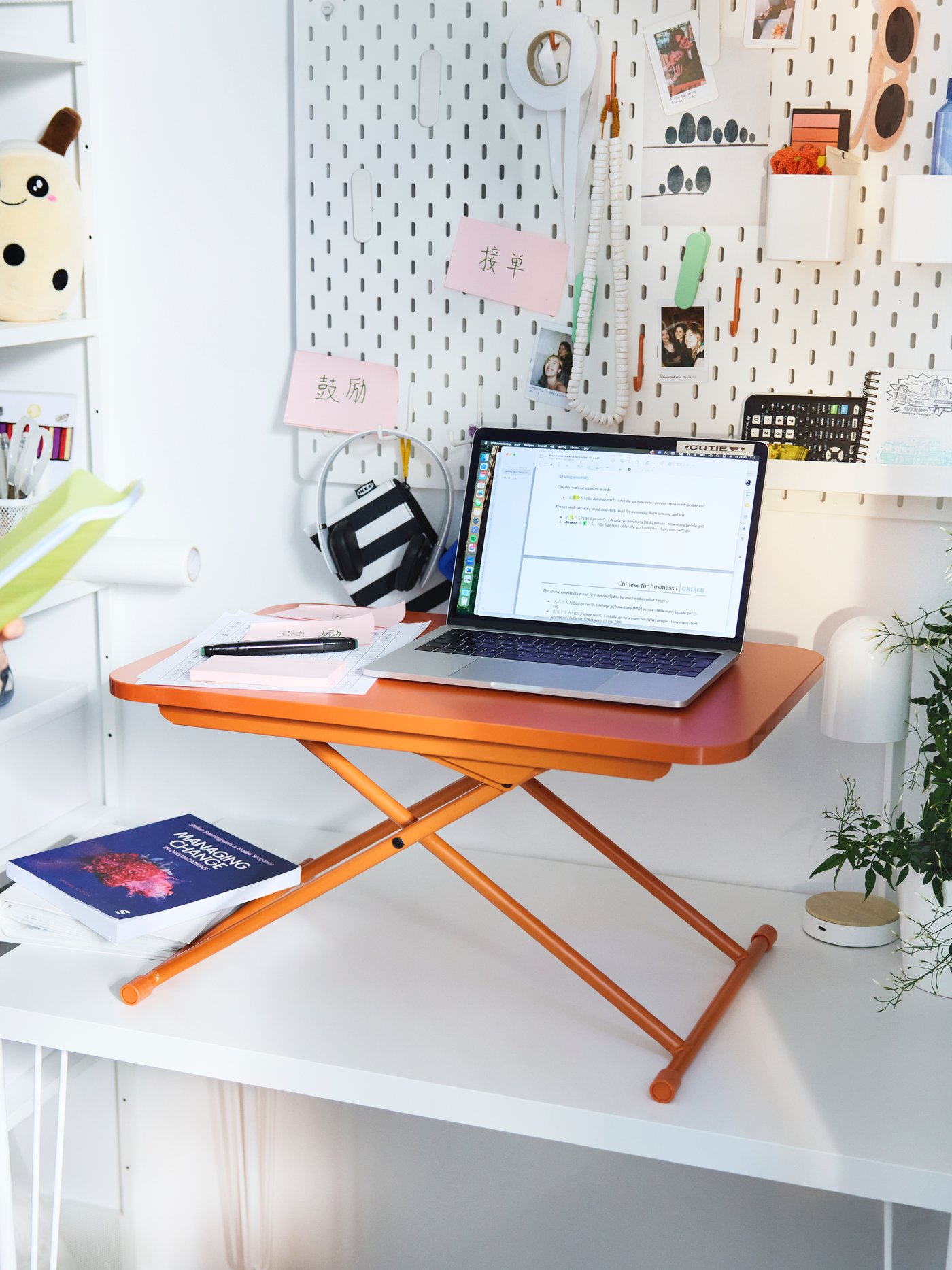 The orange DUBBLA laptop stand with a laptop placed on a white desk in front of a white pegboard and a small pink mirror.