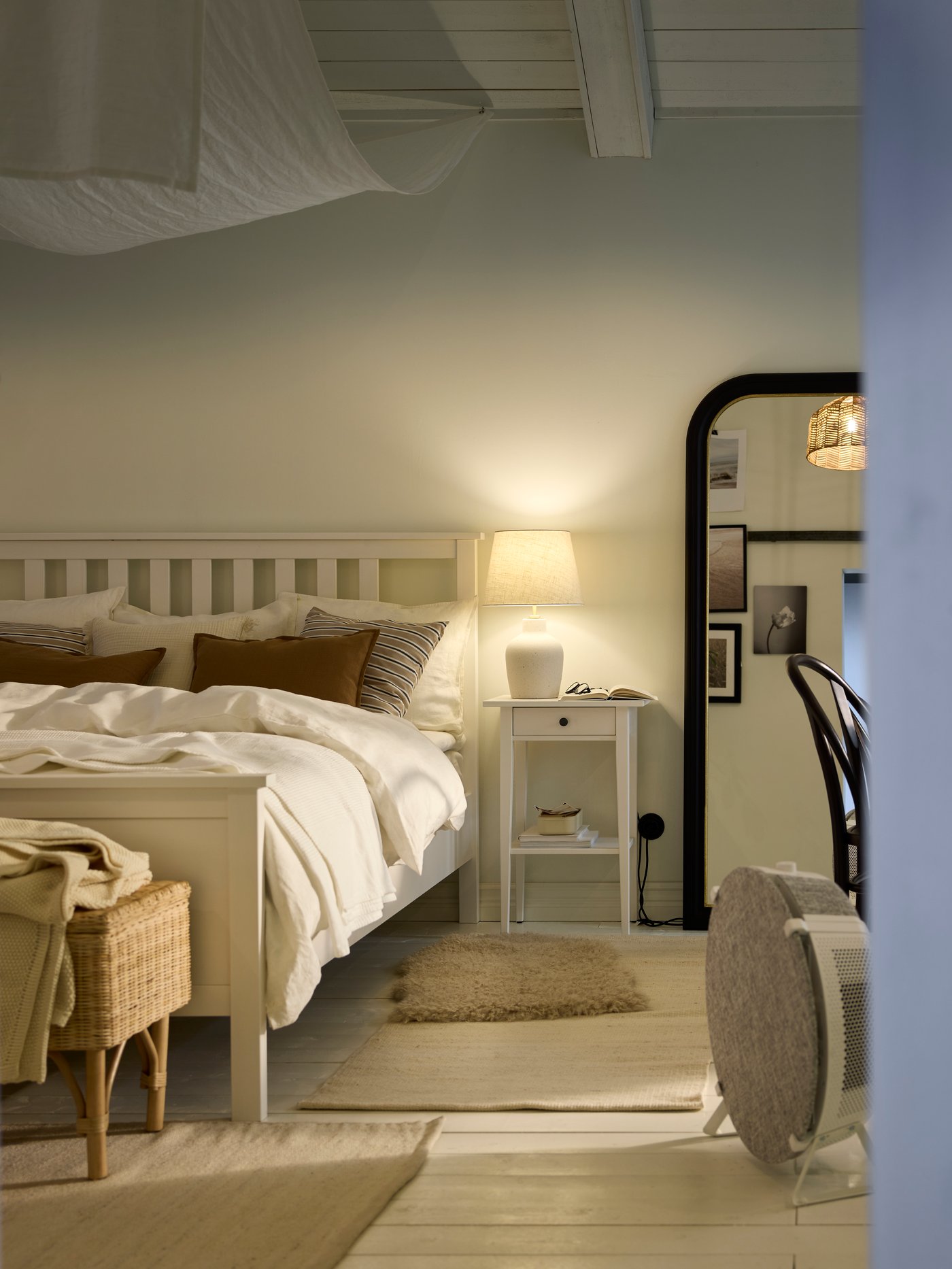 A white HEMNES bedside cabinet with a lamp on it by a white HEMNES bed. A STARKVIND air purifier is by the end of the bed.