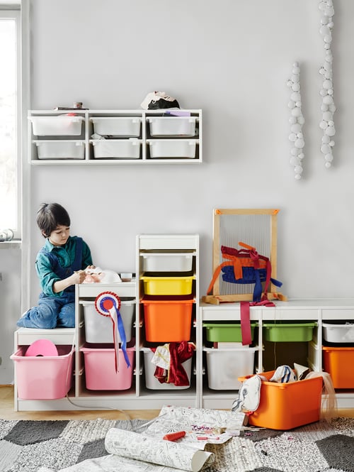 A child sits on part of a staircase-like, white TROFAST frame with white and coloured boxes, against a wall of toy storage.