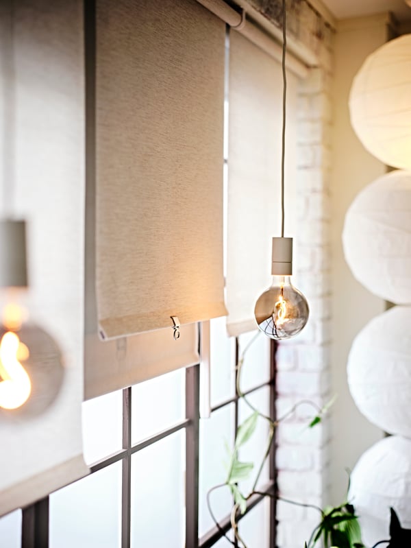 A lit MOLNART LED bulb suspended from a white SUNNEBY cord set next to a window with a beige SANDVEDEL roller blind.