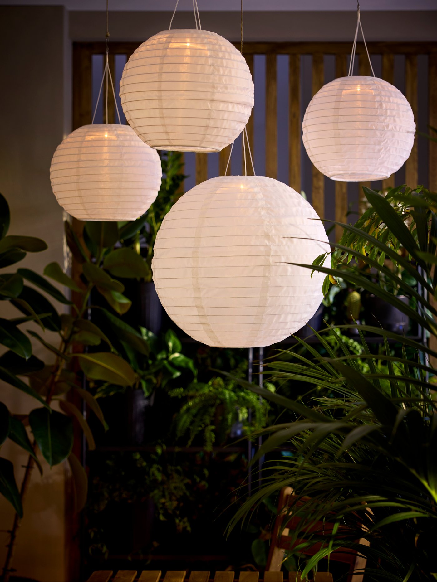 Four globe-shaped SOLVINDEN pendant lamps with white shades glow warmly as they hang from a balcony ceiling, above greenery.
