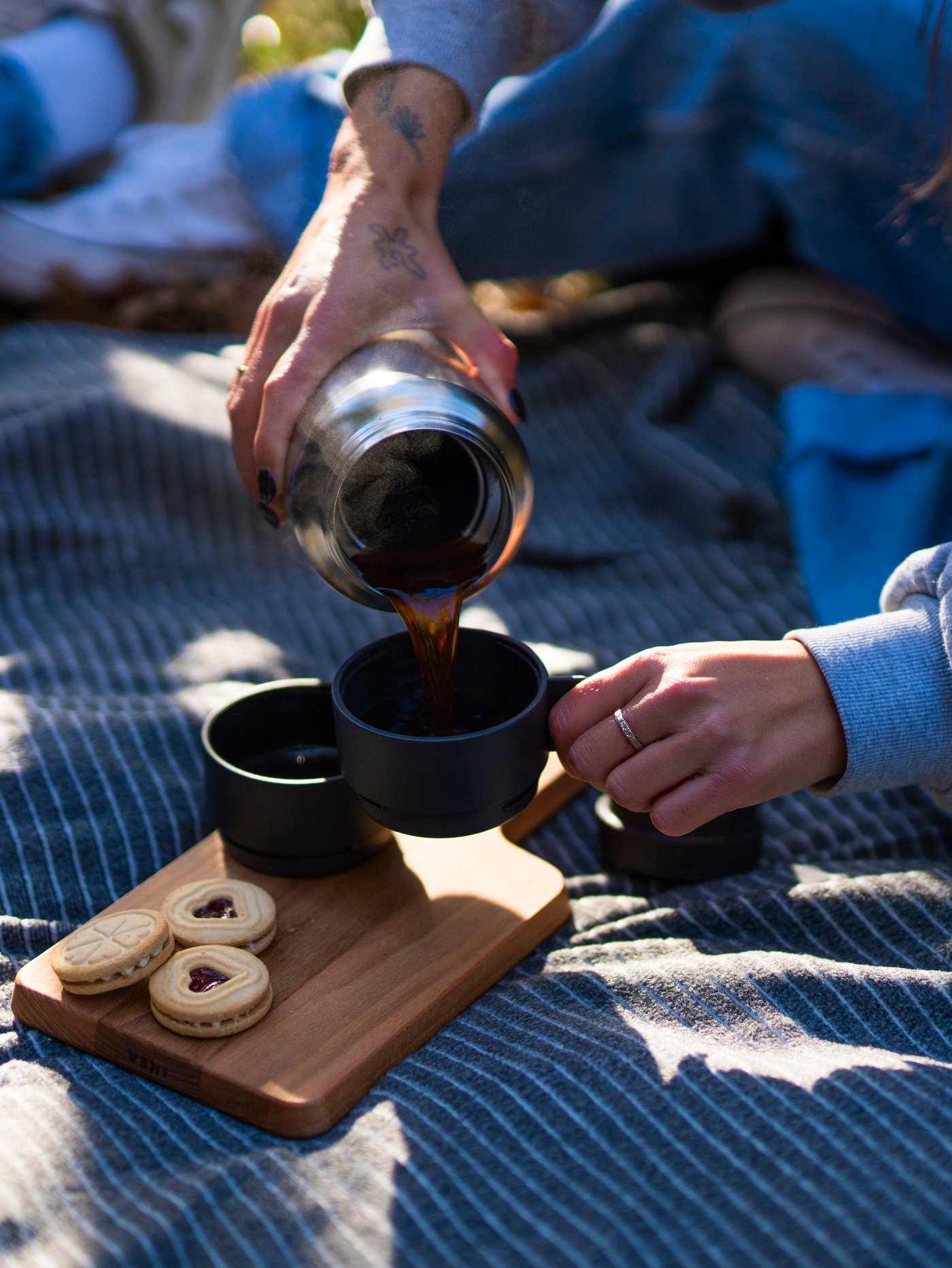 Persona vertiendo café caliente de un termo UTRUSTNING de acero en una taza pequeña, mientras está sentada en una colcha TJÄRBLOMSTER.