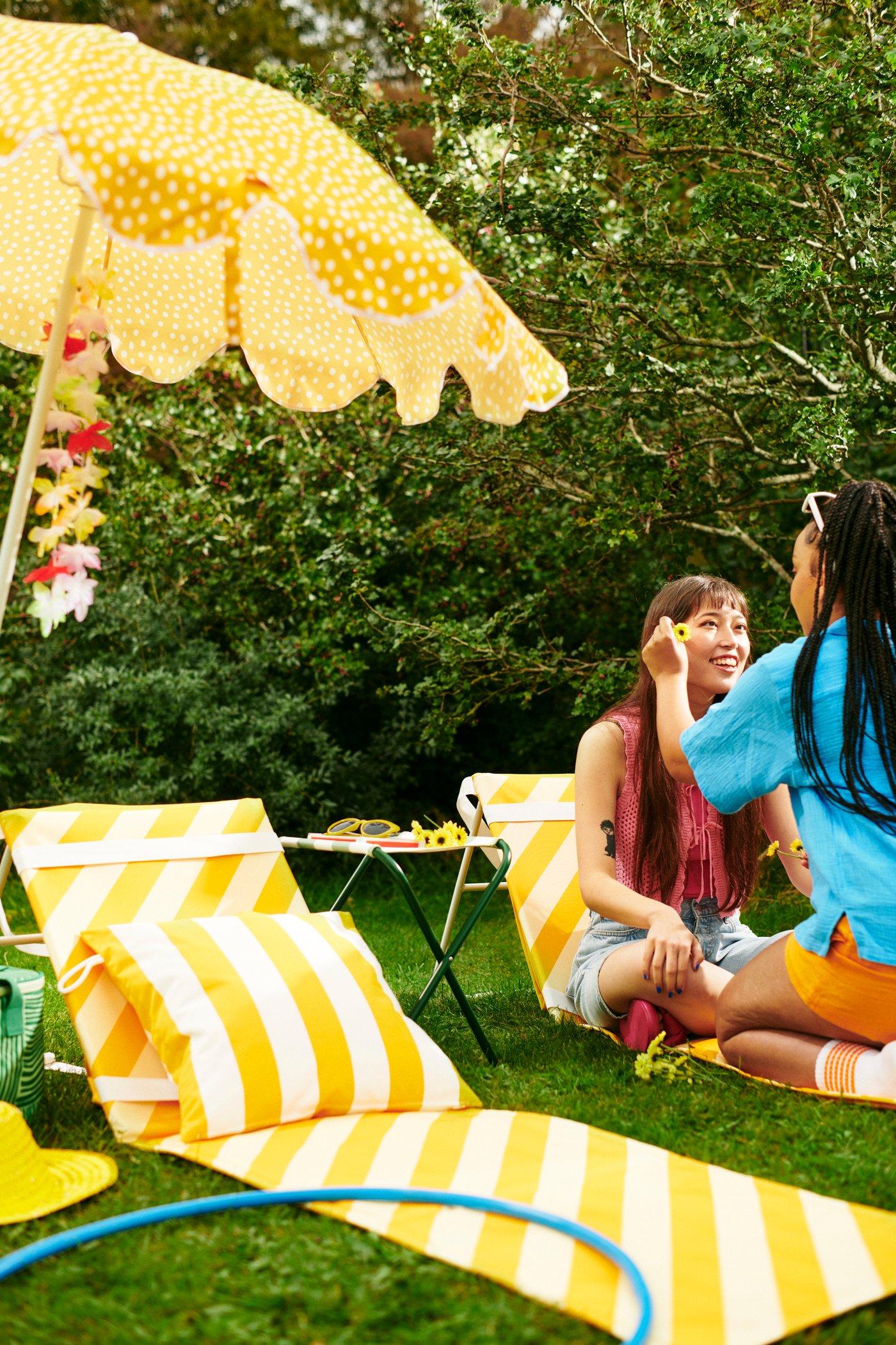 Zwei Personen sitzen auf einer Wiese neben gelb‑weiß gestreiften Liegestühlen und einem gelben Sonnenschirm. Eine Person schminkt oder bemalt das Gesicht der anderen. Im Hintergrund stehen grüne Büsche.
