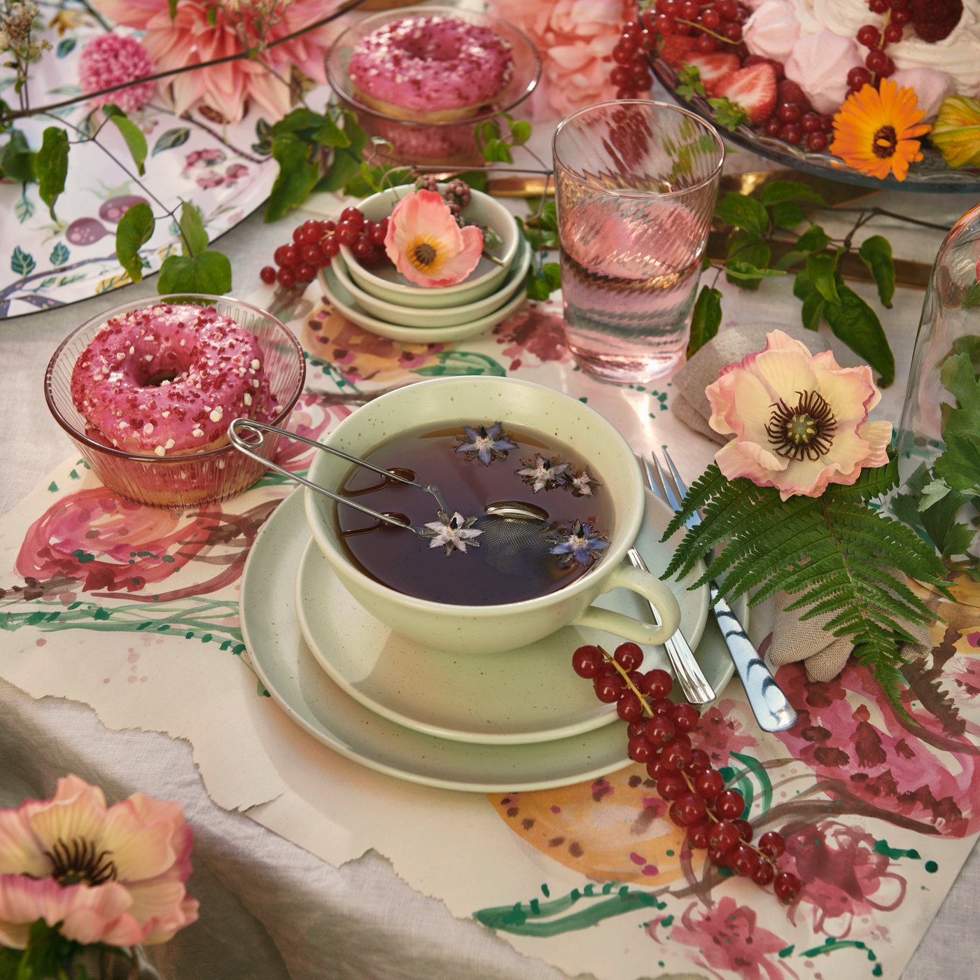 Üppig gedeckter, blütenreicher Tisch mit pastellgrüner Teetasse, in der Tee mit Sternanis schwimmt, dazu rosa Donuts, roten Beeren, Blumen und Wasserglas – eine verspielte Teepause voller Farbe.
