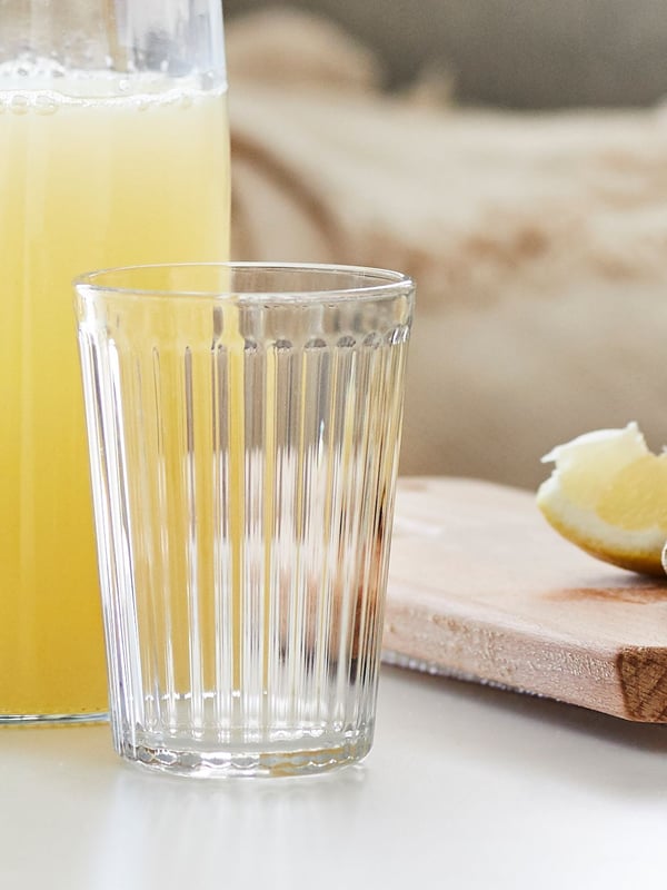 VARDAGEN Glas vor einer mit Saft gefüllten Karaffe, daneben ein Holzbrett mit einem Schnitz Zitrone
