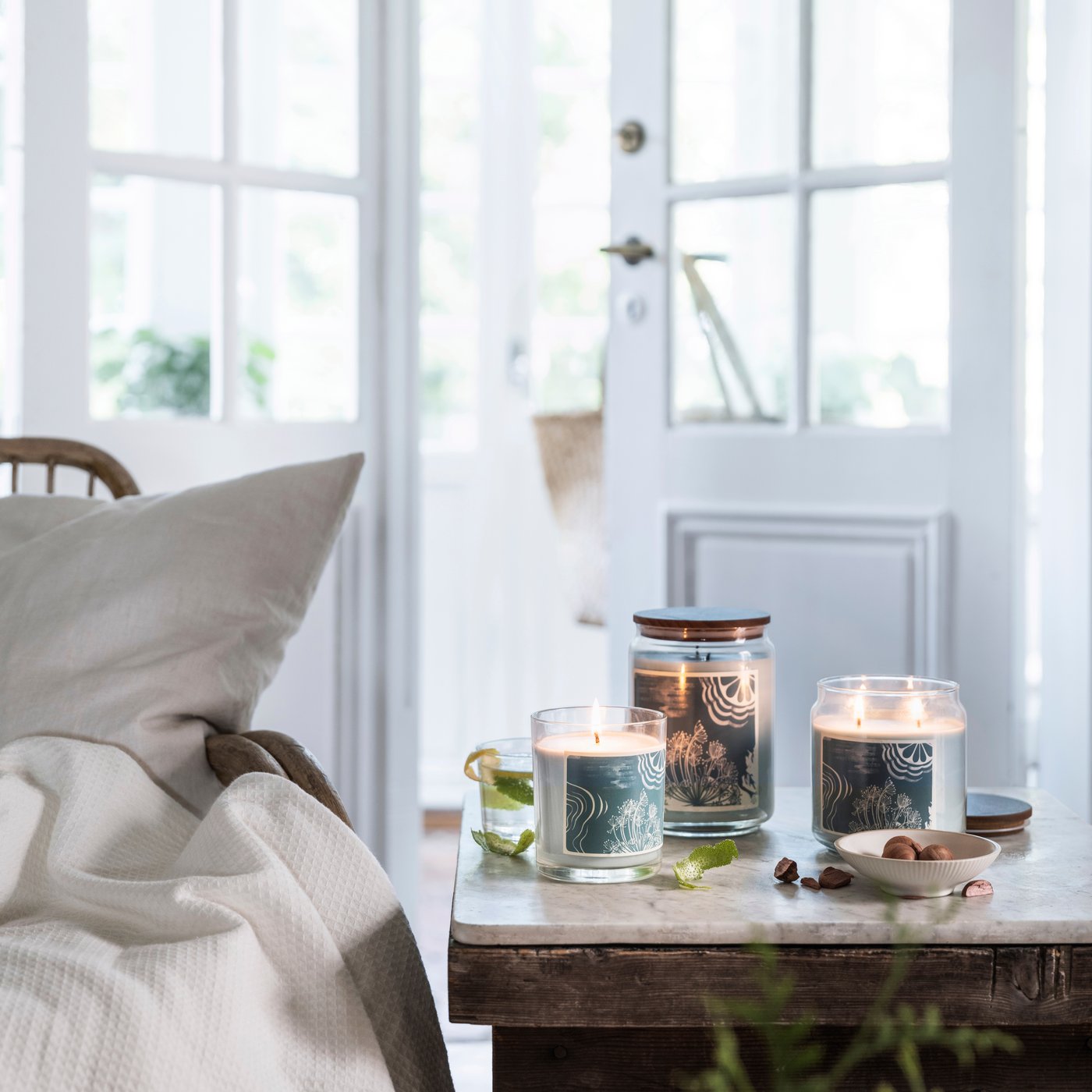 Zapálené svíčky IKEA ve skleněných dózách s dekorativními etiketami na dřevěném stole, vedle ořechů a limetkové kůry, v útulném interiéru s pohovkou a dekou.