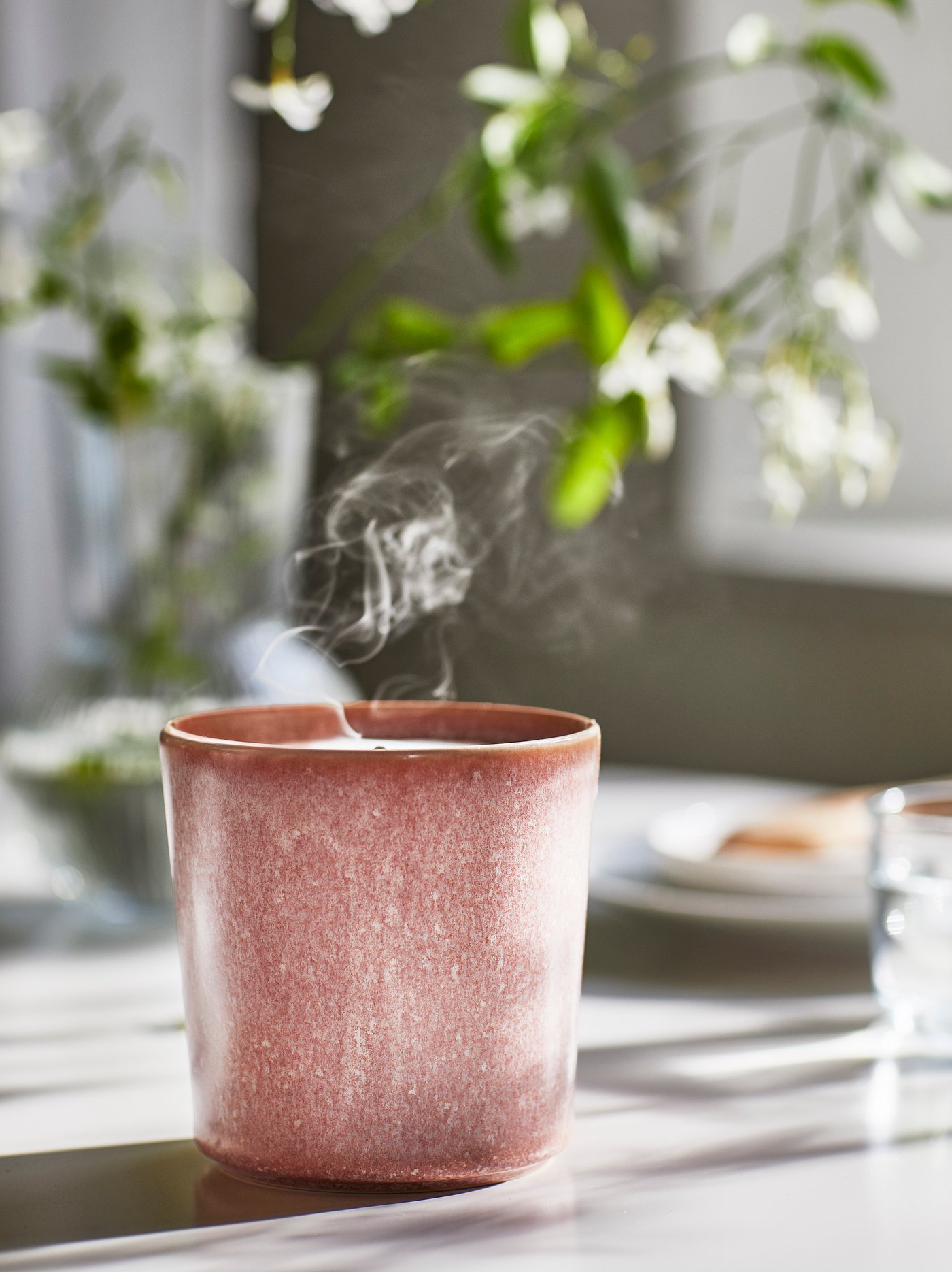 Lilin wangi LUGNARE di dalam balang seramik pada meja yang disinari cahaya matahari dan vas jelas dengan dahan bunga berwarna putih di latar belakang.