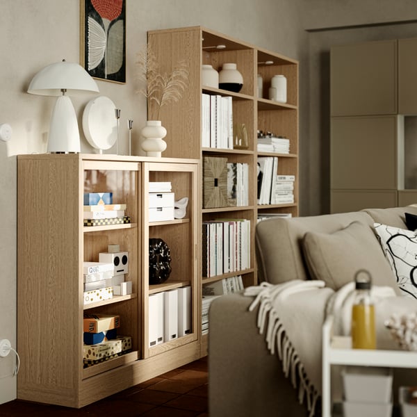 An off-white KLIPPOXEL throw lies over the arm of a sofa near an oak veneer TONSTAD cabinet with sliding glass doors.