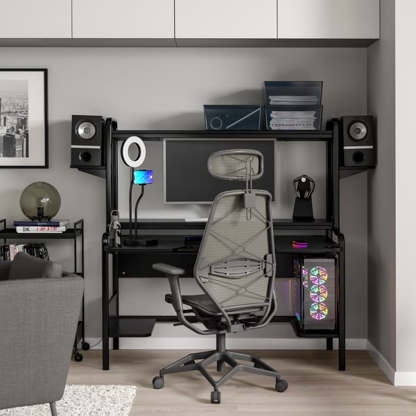 Een zwart/grijs gaming bureau en stoel met een ringlamp en wandkasten.