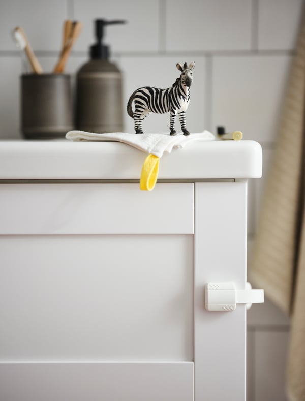 Un seguro para cajones UNDVIKA fijado en la esquina de un mueble de baño blanco. Sobre el mueble hay una cebra de juguete, una toalla de bebé KRAMA y, al fondo, accesorios de baño. 