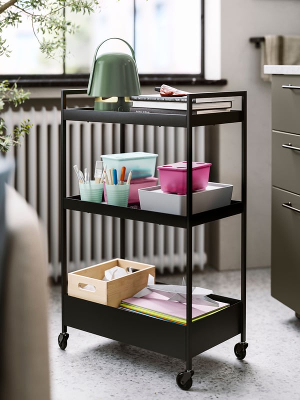 A black NISSAFORS trolley holding a green VAPPEBY Bluetooth speaker lamp, books, boxes, pens in mugs and coloured paper.