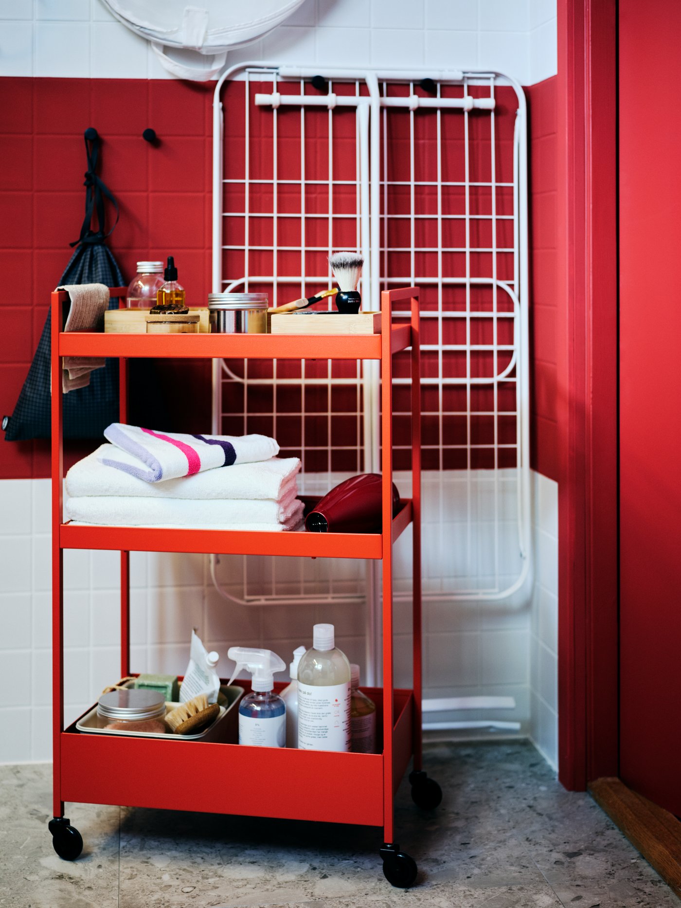 En la esquina de un cuarto de baño con azulejos rojos y blancos se encuentra un carrito NISSAFORS rojo con accesorios.