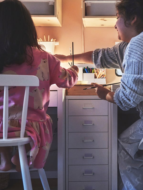En vit HELMER hurts på hjul står mellan två skrivbord där två barn sitter och pysslar.