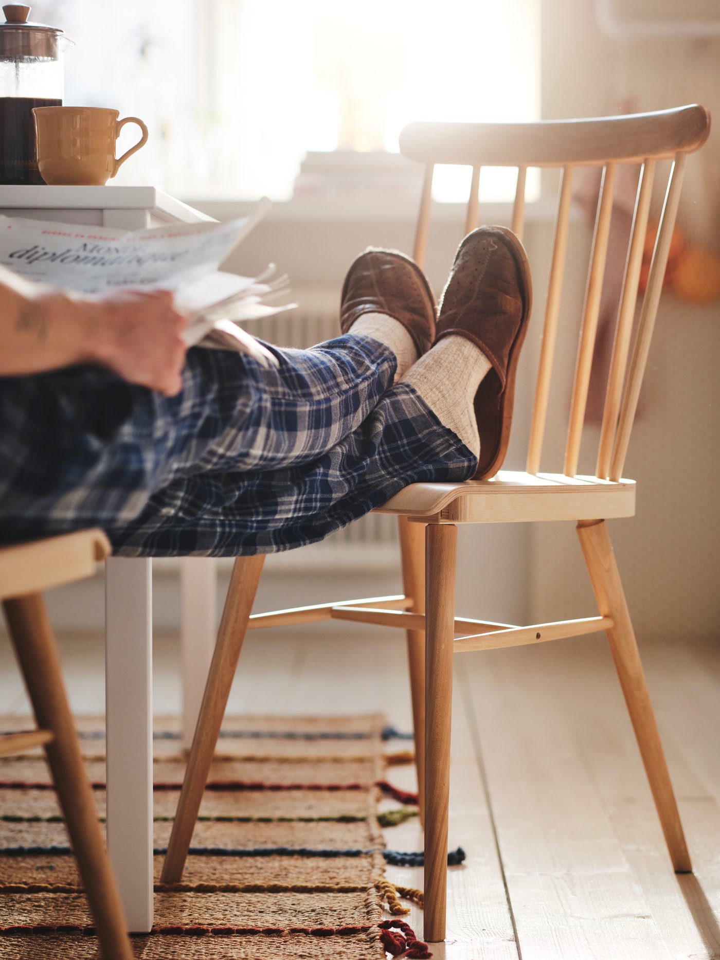 En man klädd i blårutiga pyjamasbyxor och tofflor läser tidningen i köket med fötterna upp på en matsalsstol.