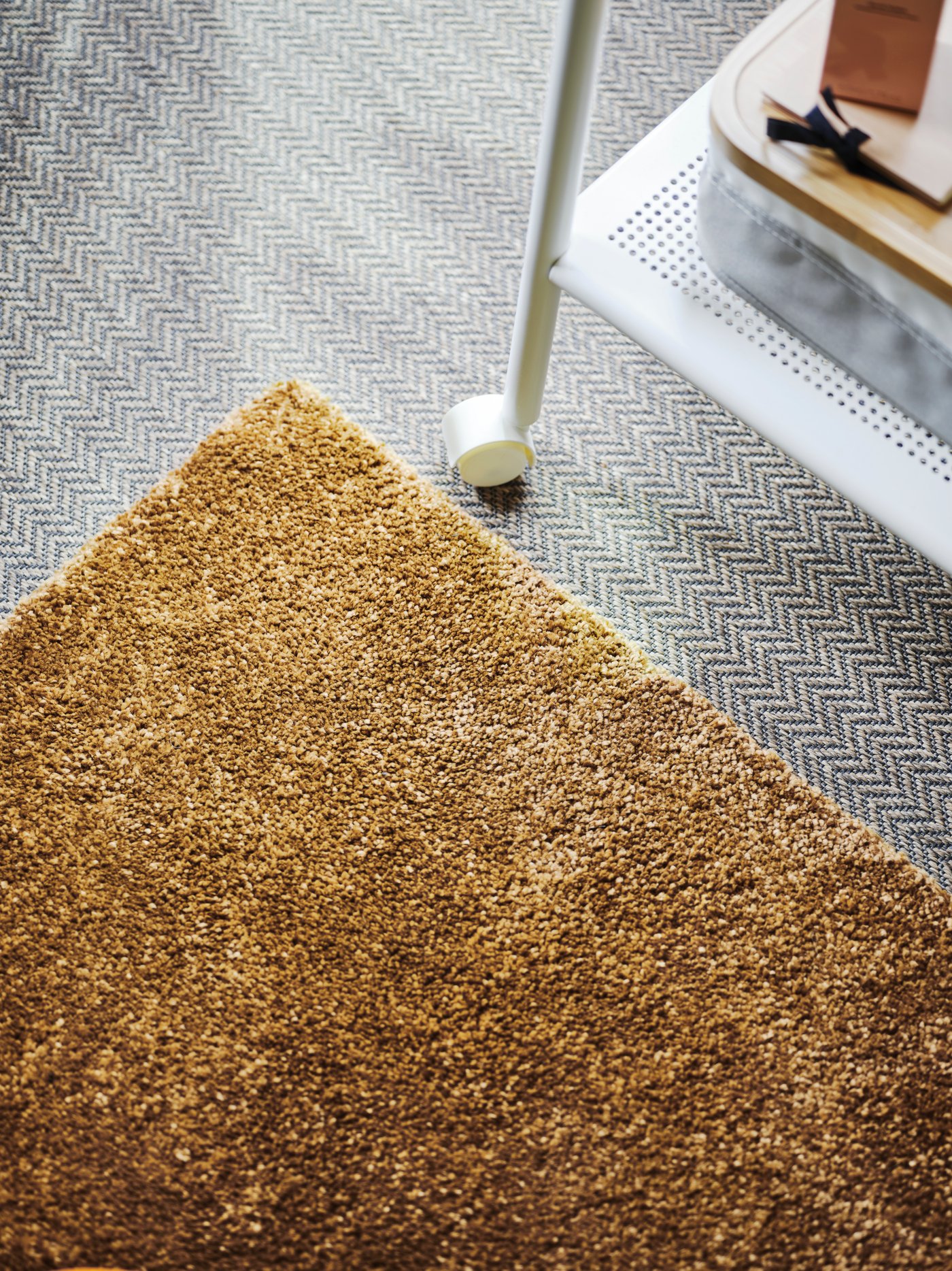 Grey flooring with a golden brown STOENSE rug with low pile and the leg and lower shelf of a white bedside table.