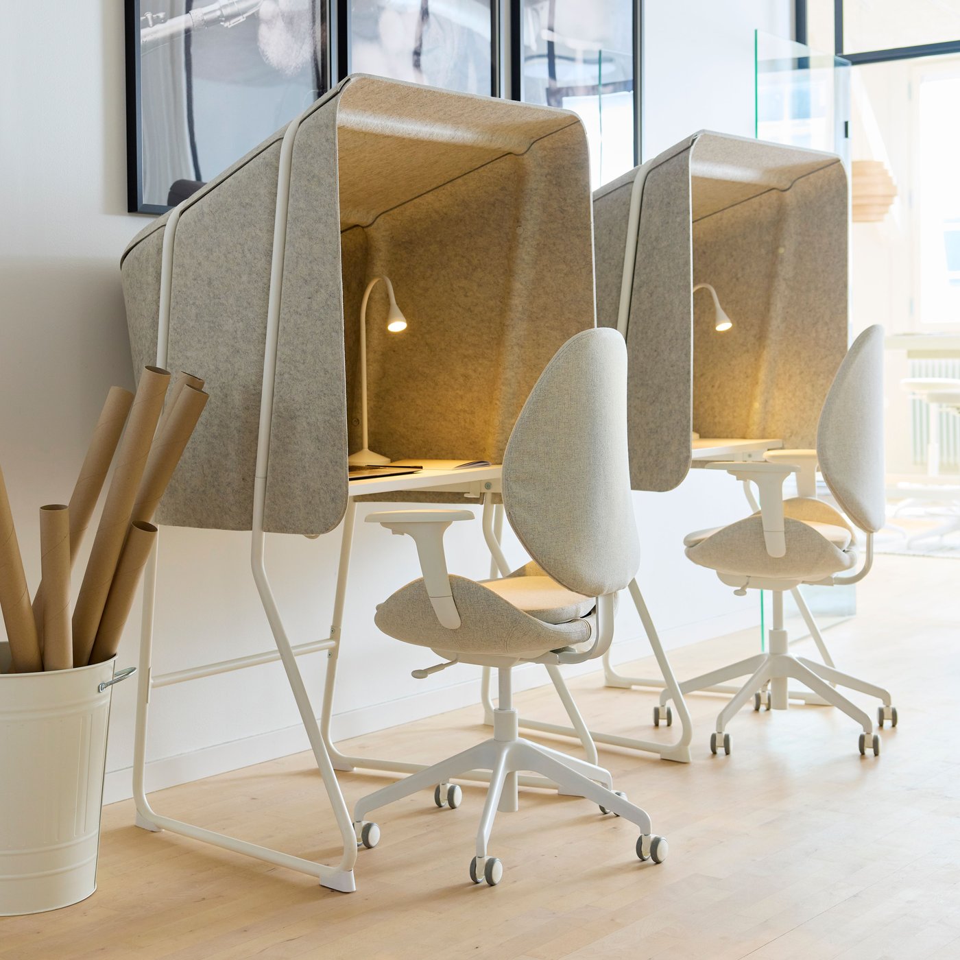 Två ljuddämpade arbetsstationer med höga tygväggar, små skrivbord och kontorsstolar på hjul. Varje plats har en integrerad lampa, och rummet har ljusa golv och stora fönster.