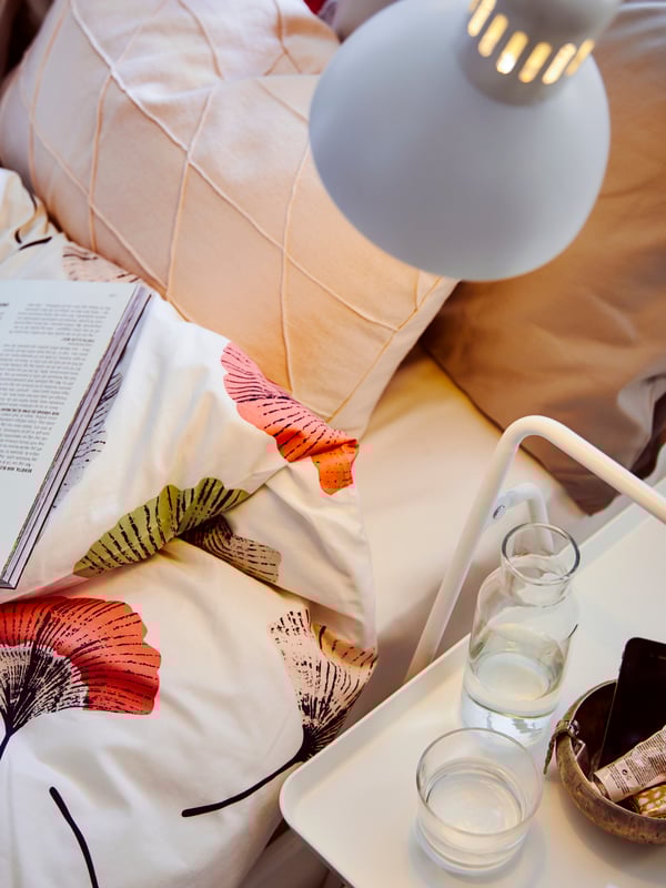 Une lampe de travail TERTIAL éclaire un lit NESTTUN avec une couette florale, des oreillers et un livre, ainsi qu’une petite table d’appoint blanche.