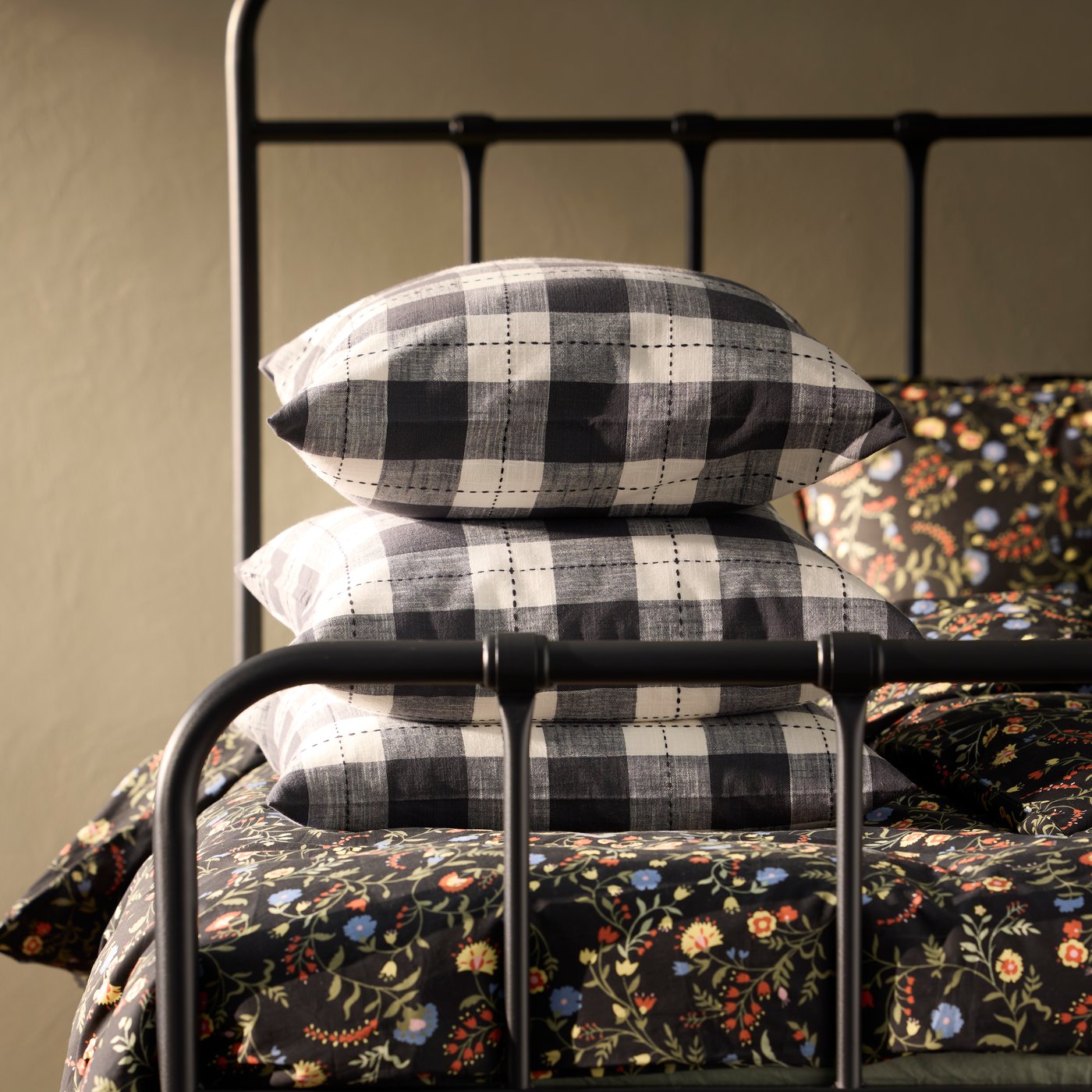 Three anthracite SVAMPFLY cushion covers with a checkered pattern are neatly placed on a floral-patterned bed.