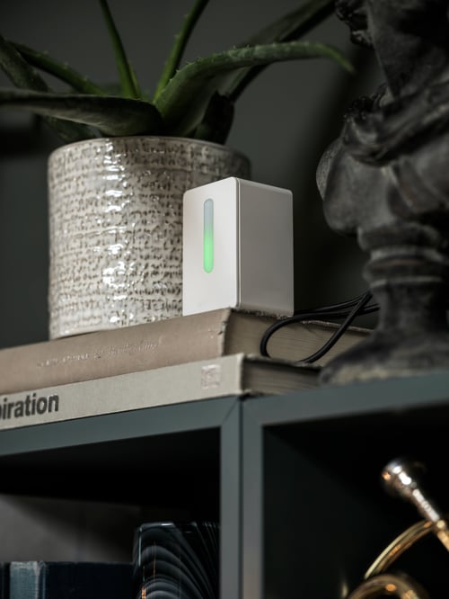 Close-up of a shelf with a white air quality sensor made out of ABS plastic placed over 2 books, next to a white plant pot.