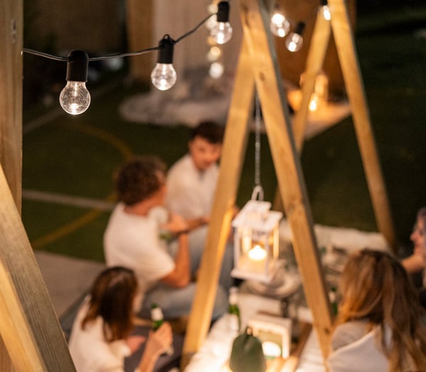 Una tavola bassa allestita e apparecchiata in uno spazio esterno illuminato da luci decorative. Attorno, persone che stanno conversando su pouf e cuscini.