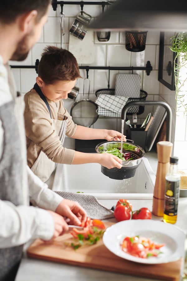 Ett barn håller ett durkslag som han använder för att skölja sallad med och en vuxen står vid sidan om och skär tomater på en bambuskärbräda.
