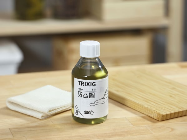A bottle of TRIXIG wood oil stands on a wooden table next to a folded tablecloth and a wooden cutting board, in a bright indoor environment.