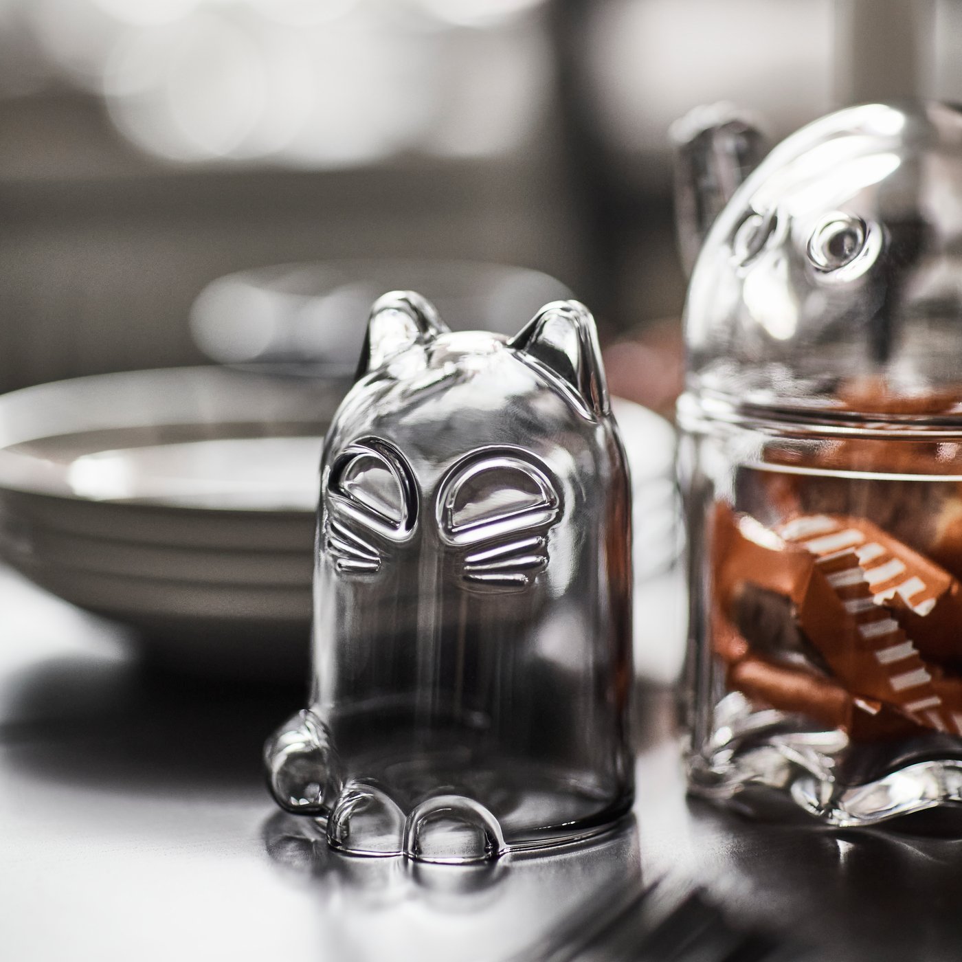 A KUSTFYR glass dome shaped like a cat is placed on a table next to a KUSTFYR bowl with lid shaped like a ghost.