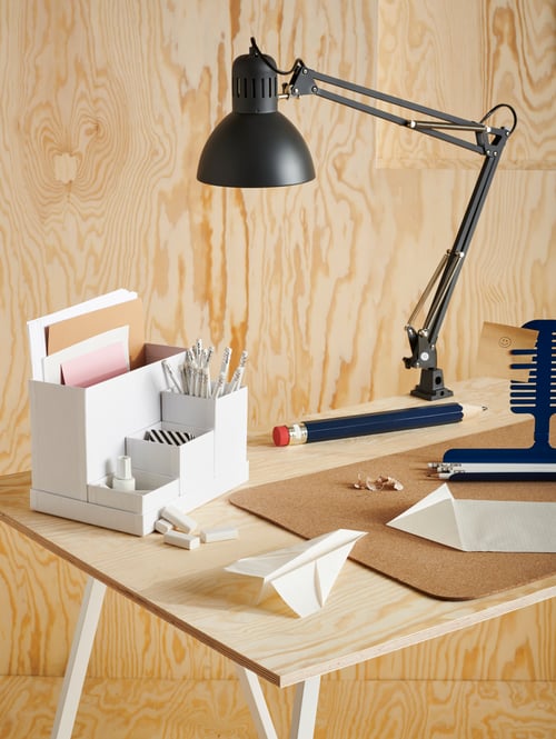 White TJENA desk organiser, MÖJLIGHET pen and picture holder, cork mat and work lamp on a wooden desk in a plywood room.