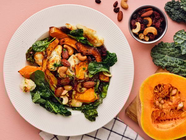 A salad with butternut squash, black kale, goat cheese, and a MUNSBIT mix of roasted nuts/berries laid out on a pink table.