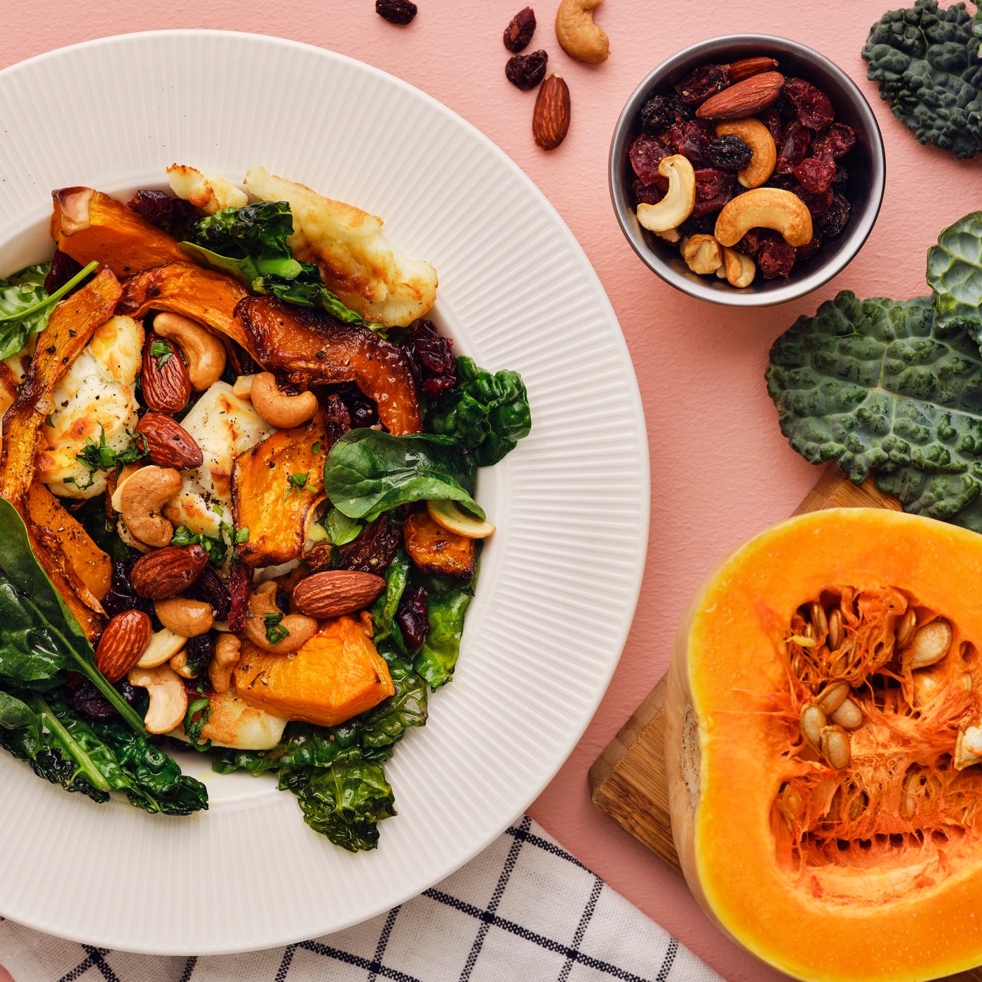 A salad with butternut squash, black kale, goat cheese, and a MUNSBIT mix of roasted nuts/berries laid out on a pink table.
