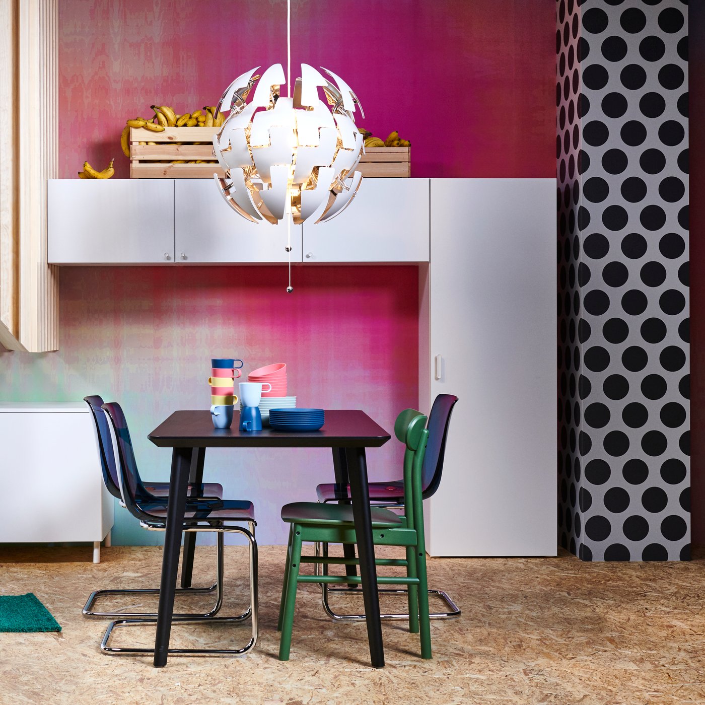 Salle à manger colorée avec une table pour quatre personnes, quatre chaises dépareillées dans une pièce avec un mur rose et des rangements muraux blancs.
