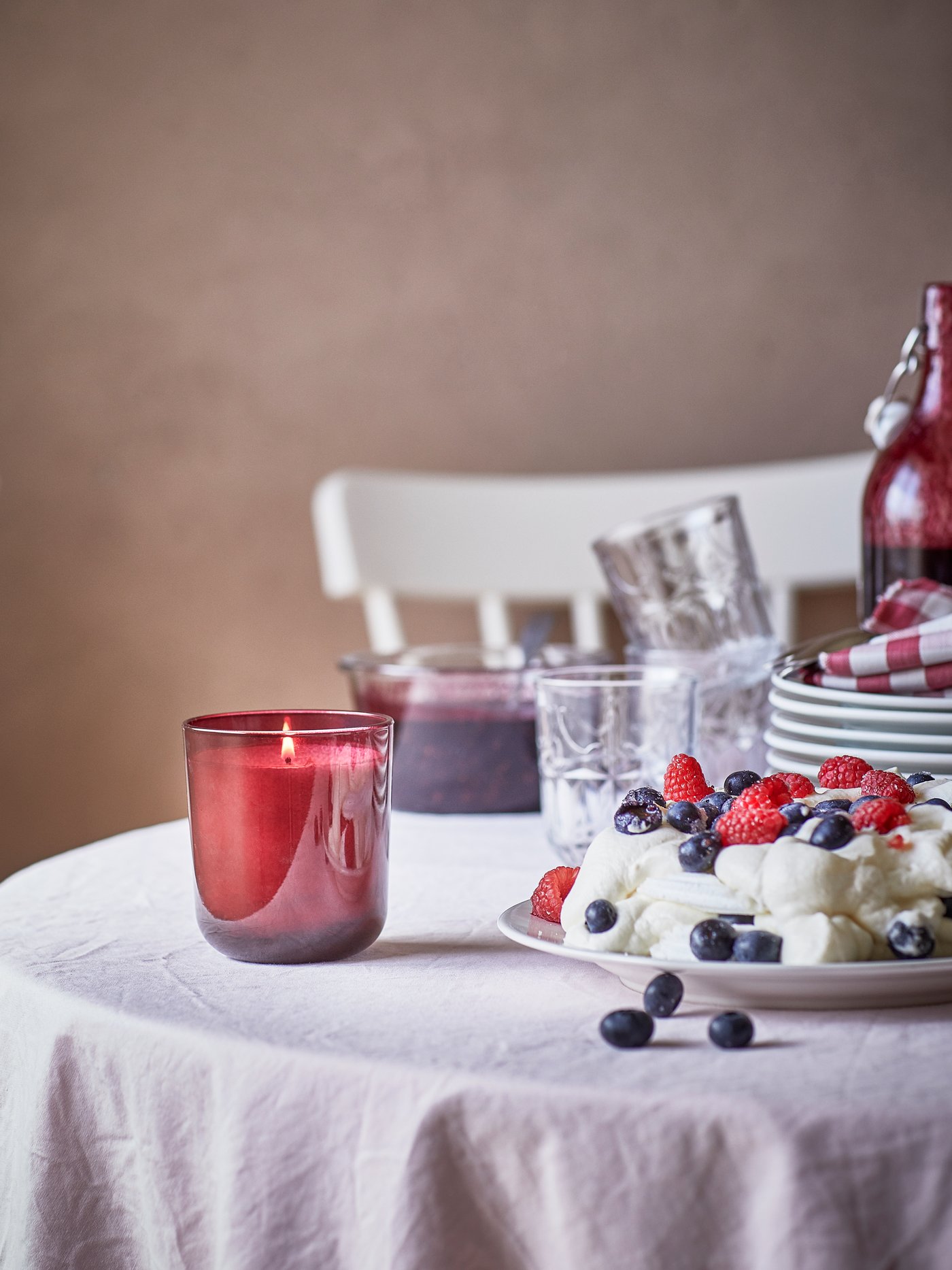 A STÖRTSKÖN scented candle in a glass jar on a table set for a celebration with a berry cake, piles of plates and glasses.